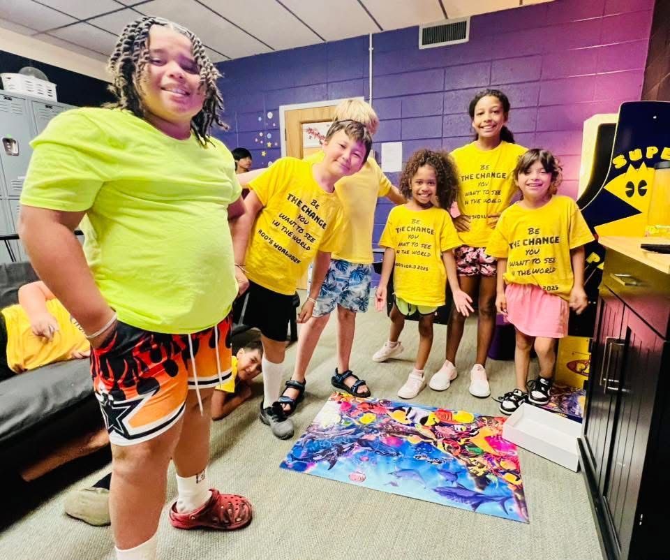 A group of children in yellow shirts gather around a colorful board game in a room with a Pac-Man arcade cabinet.