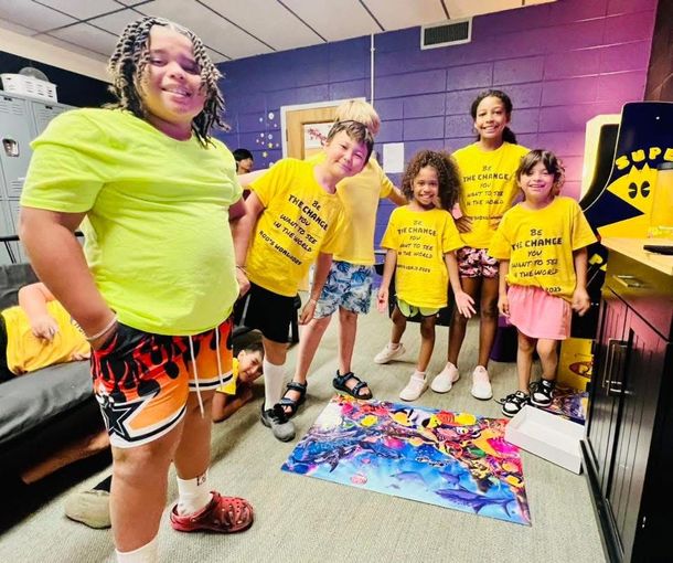 Group of people in masks, some wearing yellow shirts, smiling at the camera in a brightly lit room.