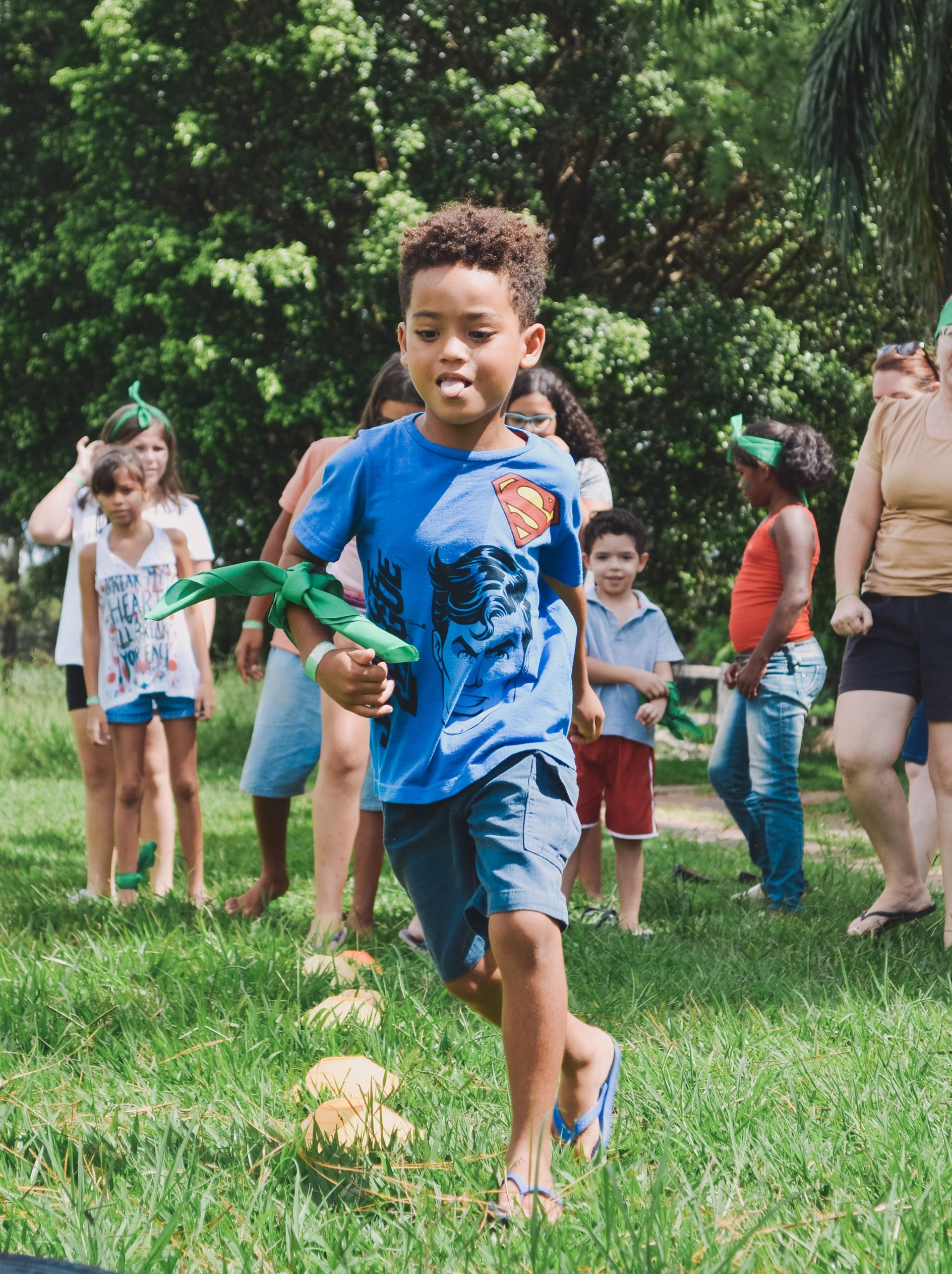 Boy running with a toy, other children and adults watching in a grassy outdoor area.