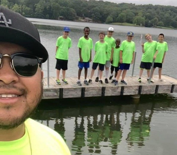 A selfie of a person in a baseball cap and sunglasses standing with a group of eight people on a wooden dock by a lake.
