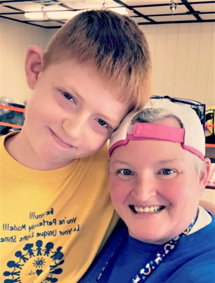 A person in a yellow t-shirt and another person in a blue shirt and pink cap lean their heads together, both smiling.