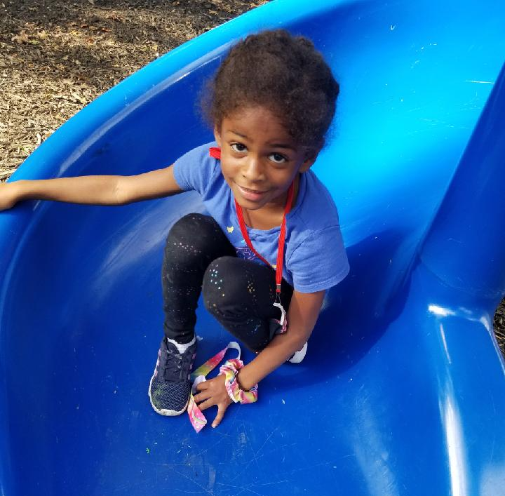 A young person sits on a blue playground slide, wearing a blue shirt and dark leggings, looking toward the camera.