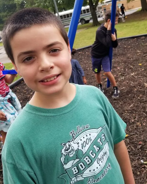A child smiles at the camera while standing on a playground with other children playing in the background.