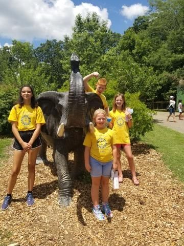 Five children in yellow shirts pose near a stone elephant statue outside on a sunny day.
