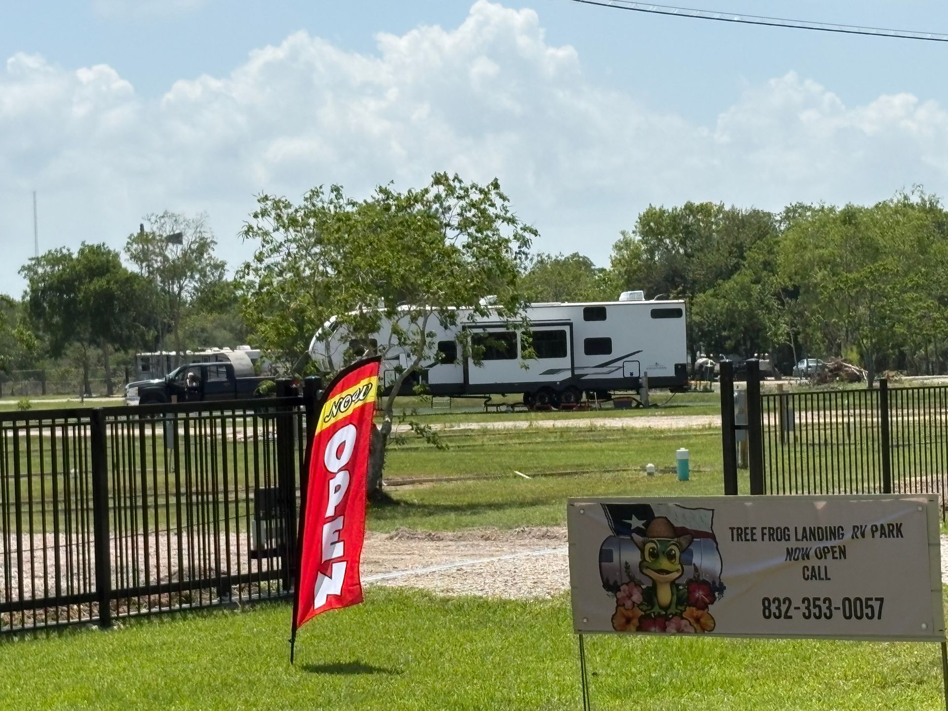 Red banner and sign by a black fence, with a white RV parked on grass in the background.