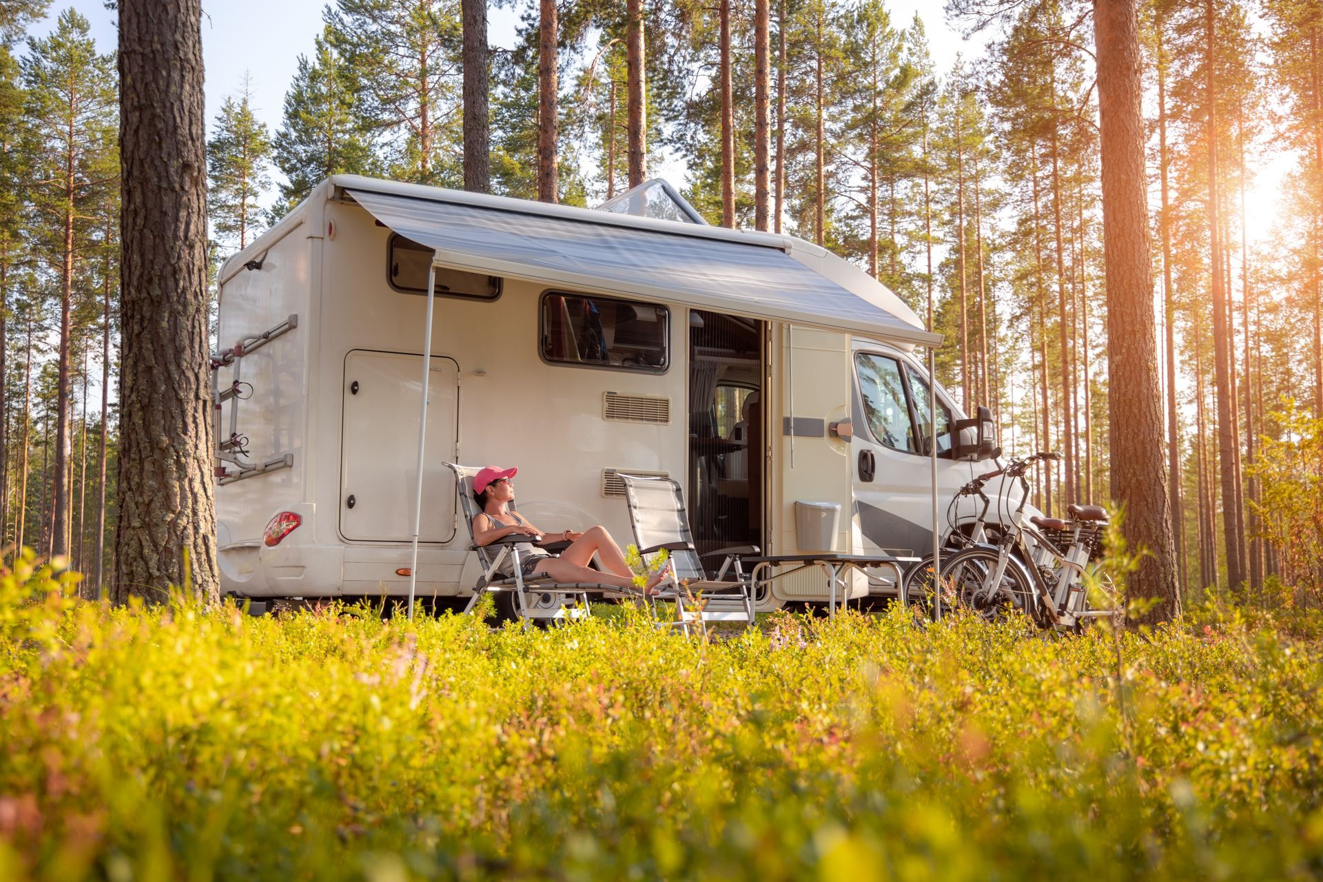 Camper van in a pine forest with a person relaxing outside in warm sunlight