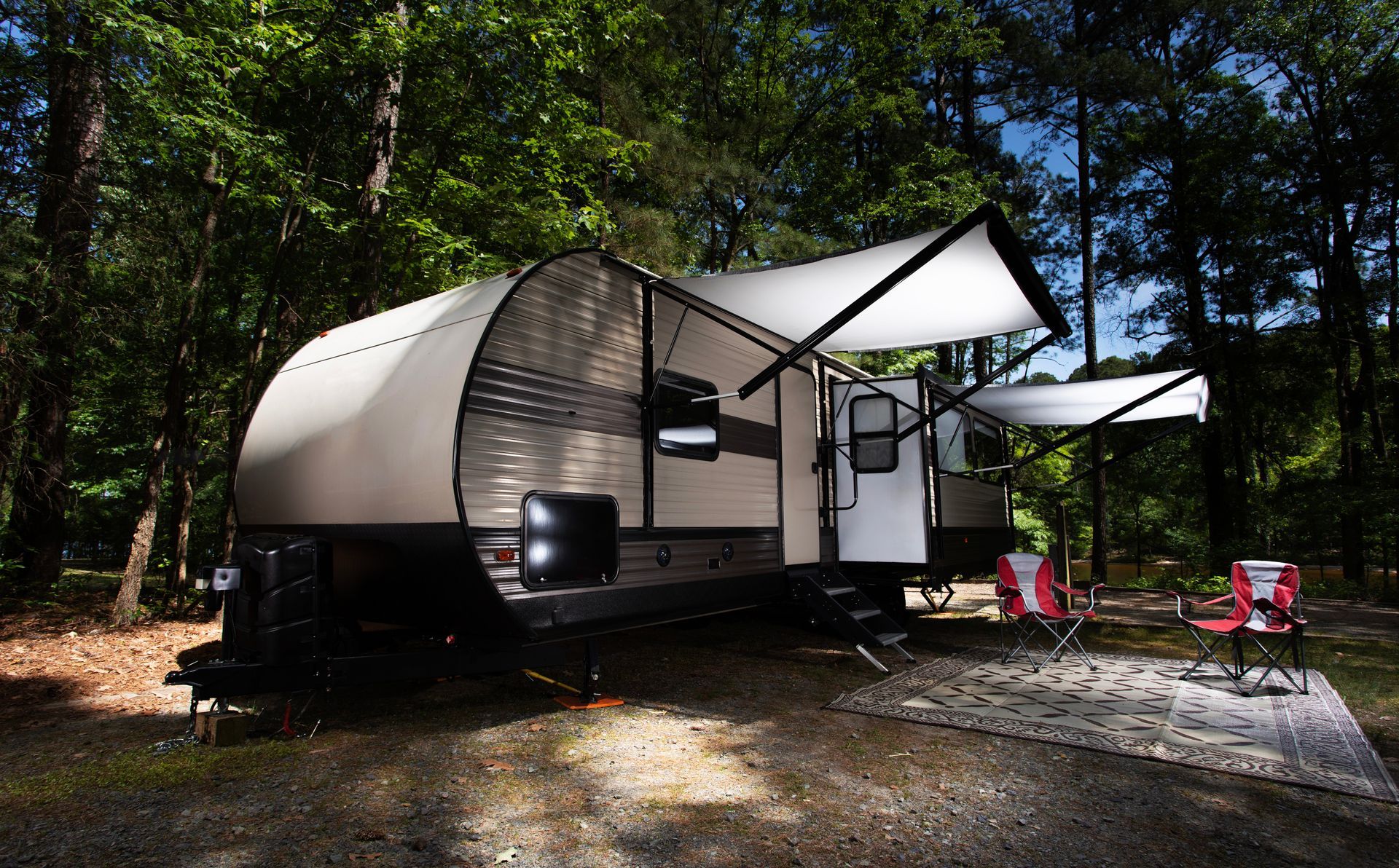 Modern travel trailer with awning in a wooded campground, with red chairs and picnic table on gravel.