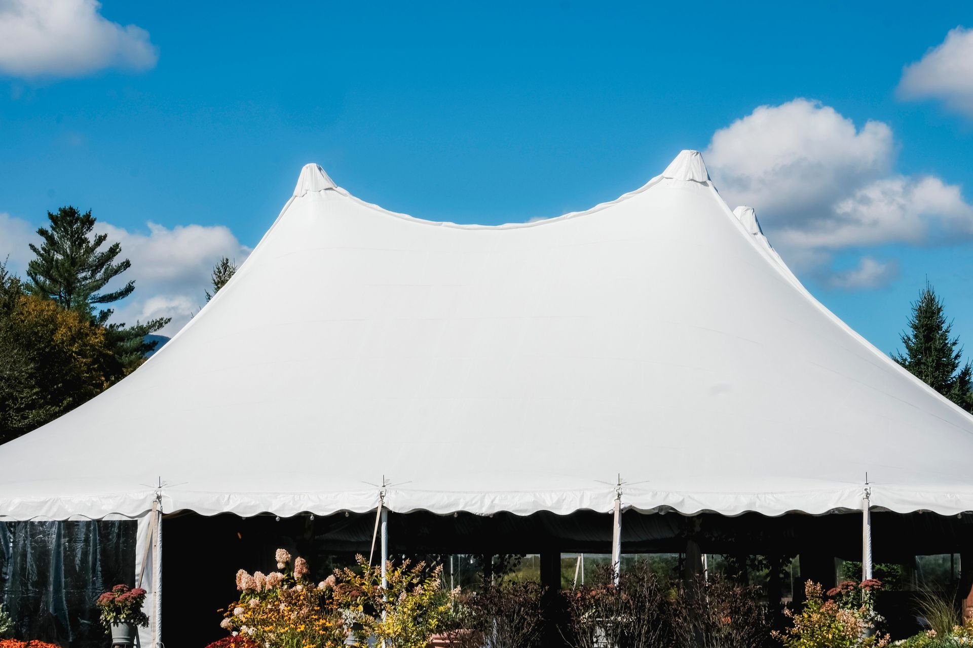 Large white event tent with peaked roof against a blue sky, surrounded by greenery and flowers
