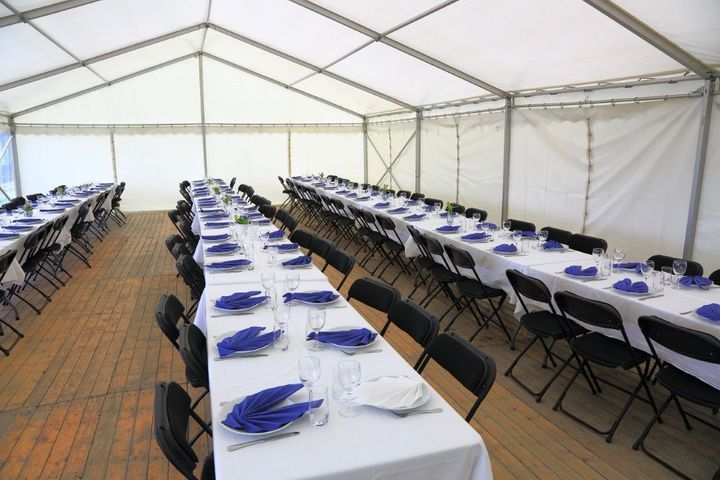 Long banquet tables set inside a white event tent, with blue napkins and folding chairs.