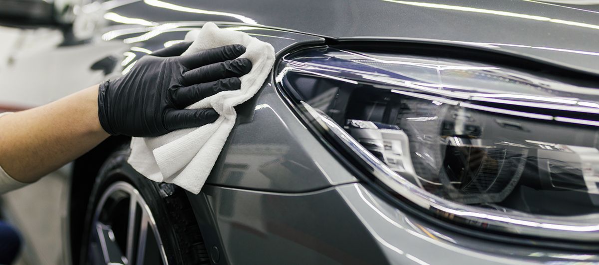 Gloved hand wiping the gray body of a car with a white cloth. Close up of the headlight.