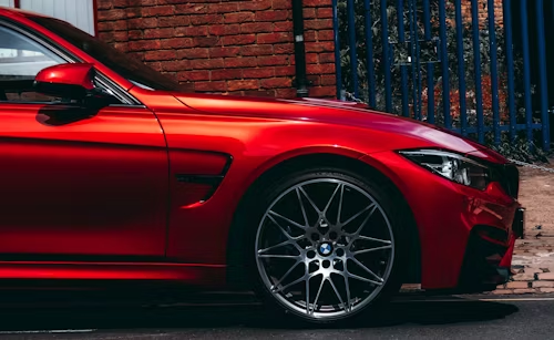 Close-up side view of a red BMW sports car parked on a street next to a brick wall and a blue metal fence.