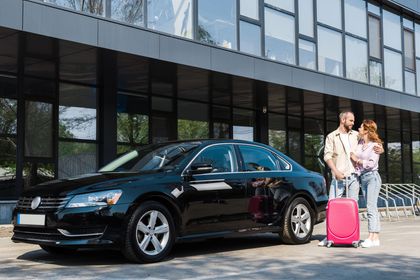 Una berlina nera parcheggiata accanto a un edificio moderno, con due persone e una valigia rosa a lato.