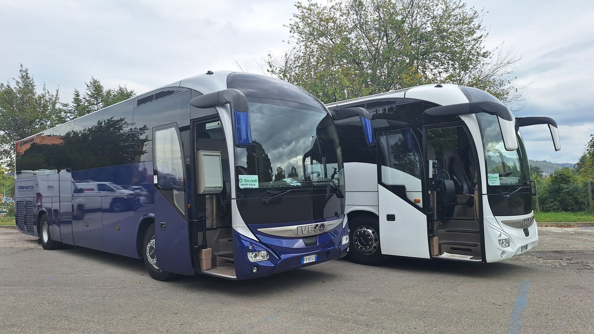 Due pullman parcheggiati, uno blu scuro e uno bianco, con le porte aperte in un parcheggio ghiaioso sotto un cielo nuvoloso