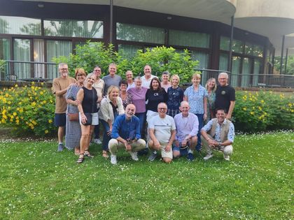 Gruppo di persone in posa su un prato di fronte a un edificio, con fiori e alberi sullo sfondo.