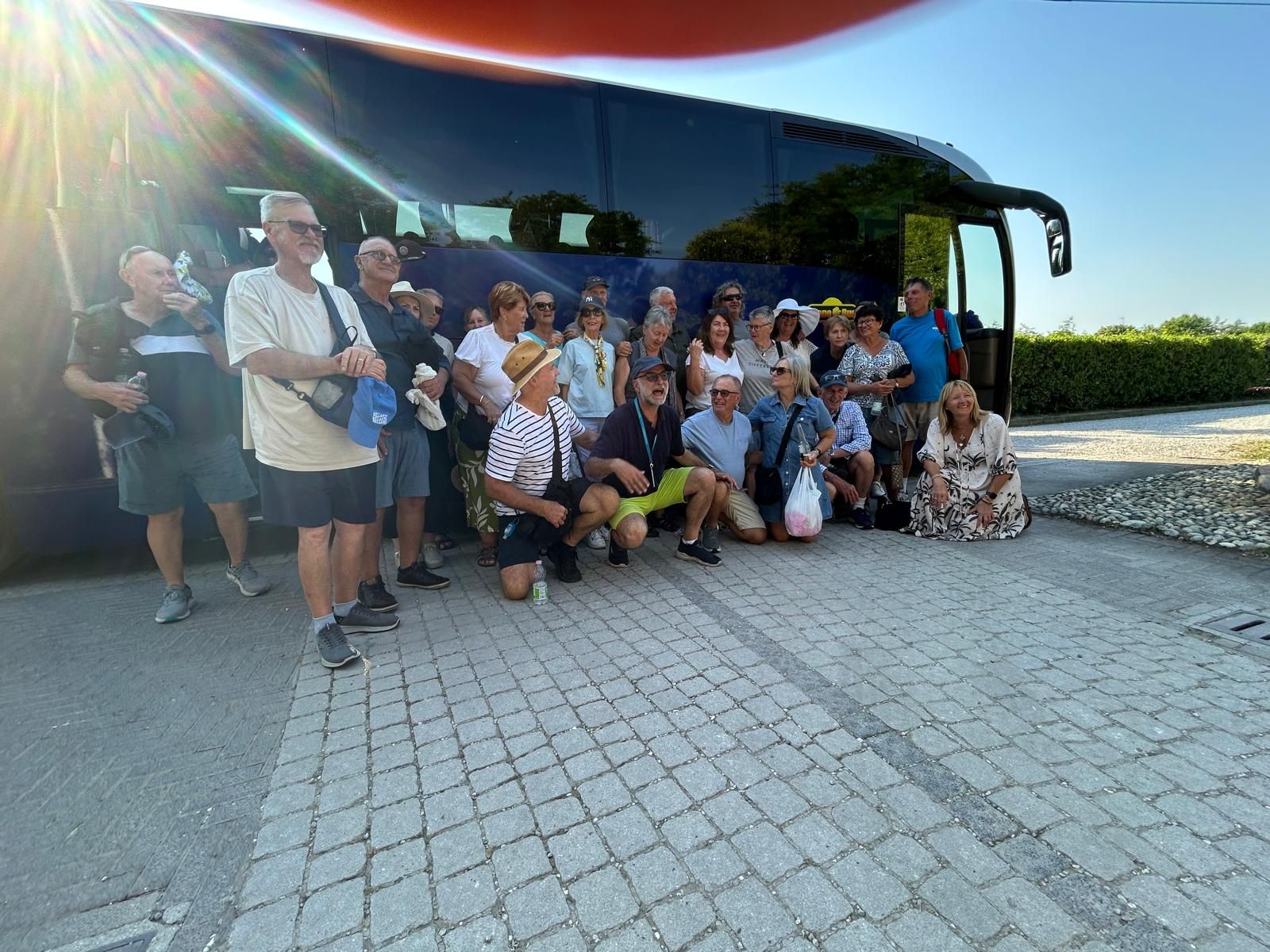 Un folto gruppo di persone posa davanti a un autobus turistico blu in una piazza asfaltata e soleggiata.