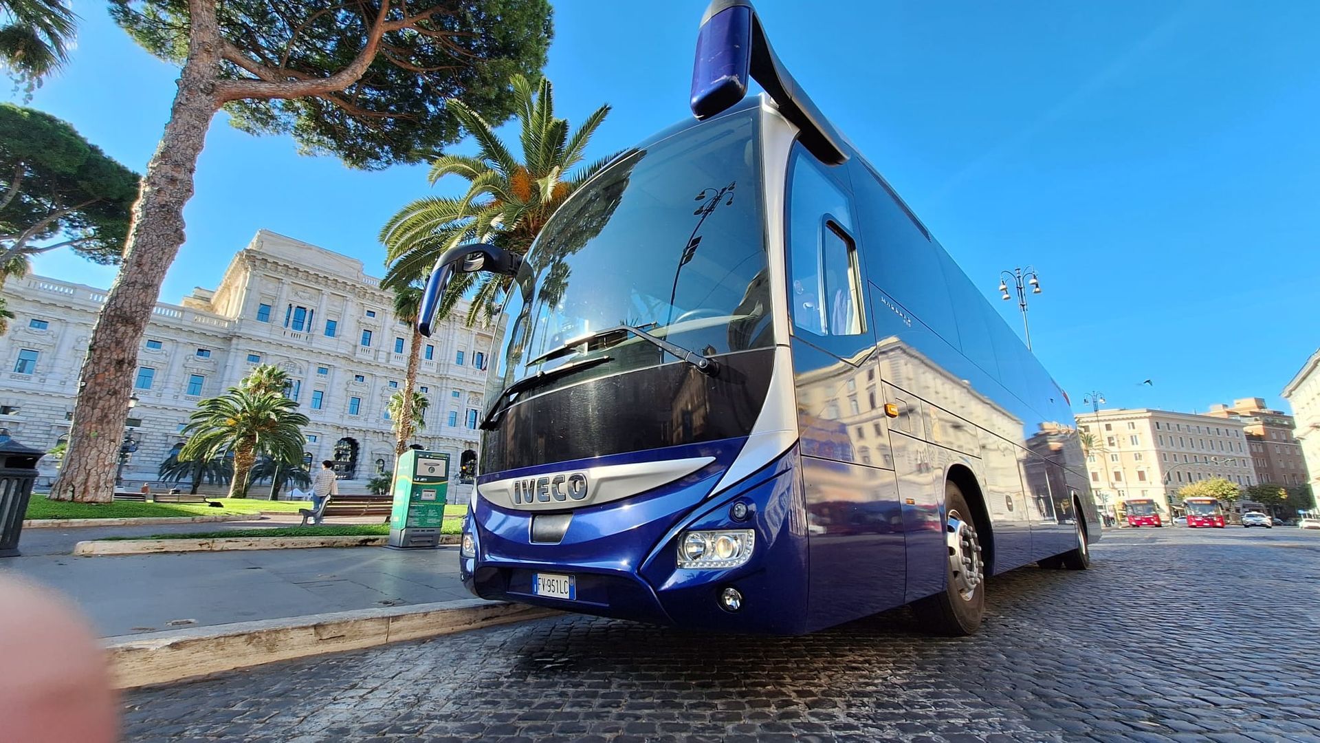 Autobus blu su una strada acciottolata in una piazza soleggiata della città, circondata da edifici storici e palme.