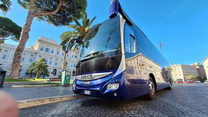 Autobus blu su una strada acciottolata in una piazza soleggiata della città, circondata da edifici storici e palme.