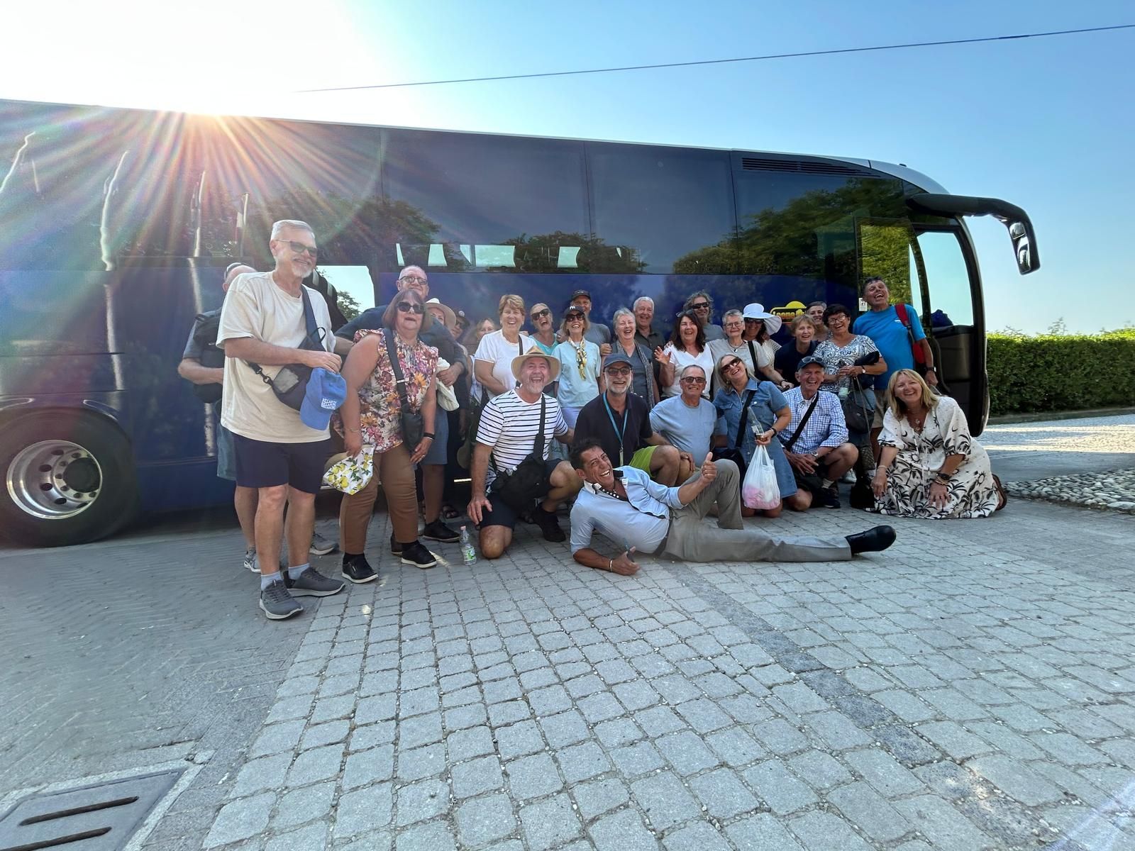 Gruppo in posa accanto a un grande autobus in pieno sole su un parcheggio asfaltato.