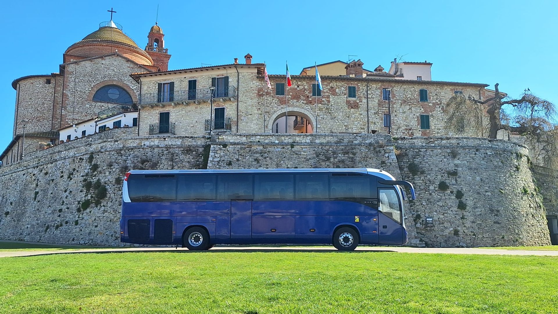 Autobus blu parcheggiato sull'erba davanti a una fortezza di pietra sotto un cielo limpido.