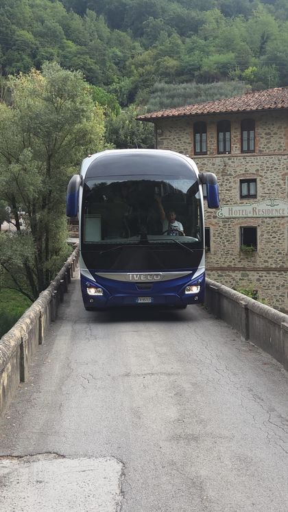 Un autobus blu su uno stretto ponte di pietra in un villaggio rurale con alberi e una casa in pietra nelle vicinanze.