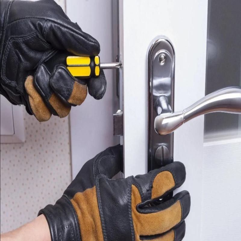 Hands in black and tan work gloves use a yellow-handled screwdriver on a white door near a metal handle.
