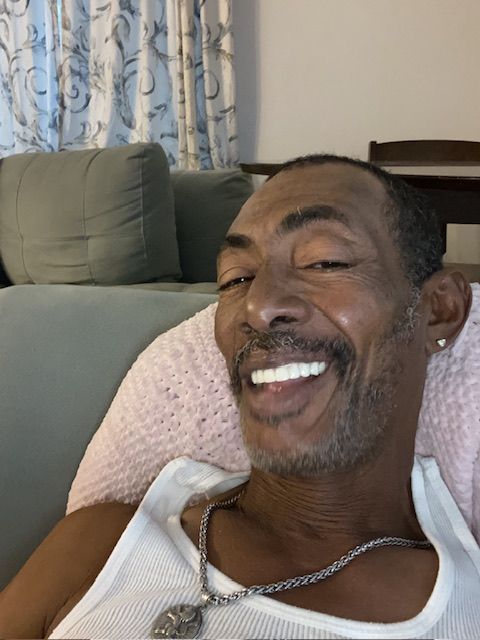 Man smiling, sitting on a couch, wearing a white tank top and silver chain.