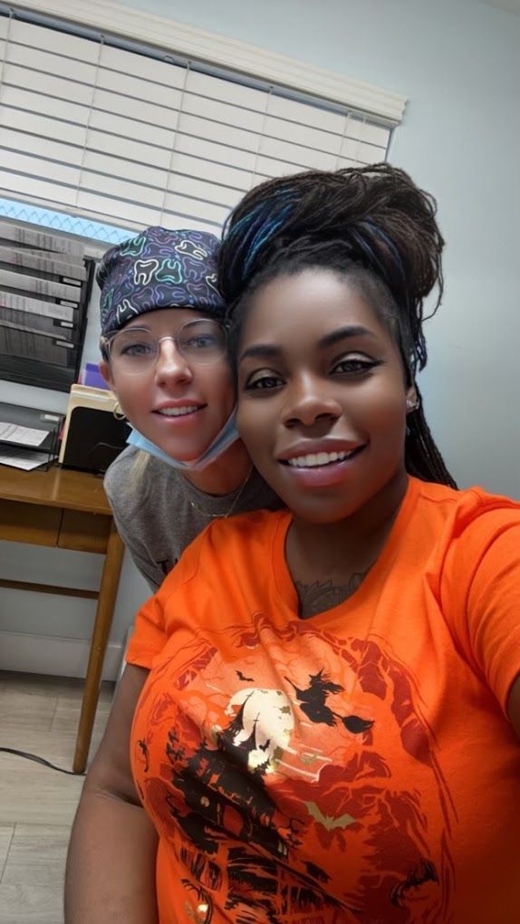 Two people smile at the camera. One wears an orange Halloween shirt, the other a patterned surgical cap. Indoor setting.