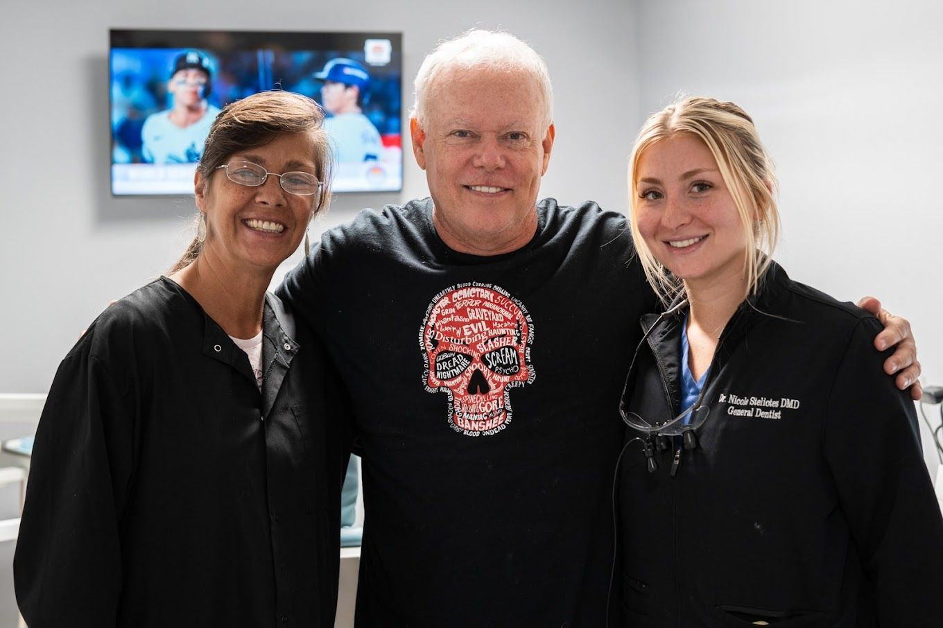 Man with two dental staff, smiling, arms around each other. Black tops, skull shirt. Dental office setting.
