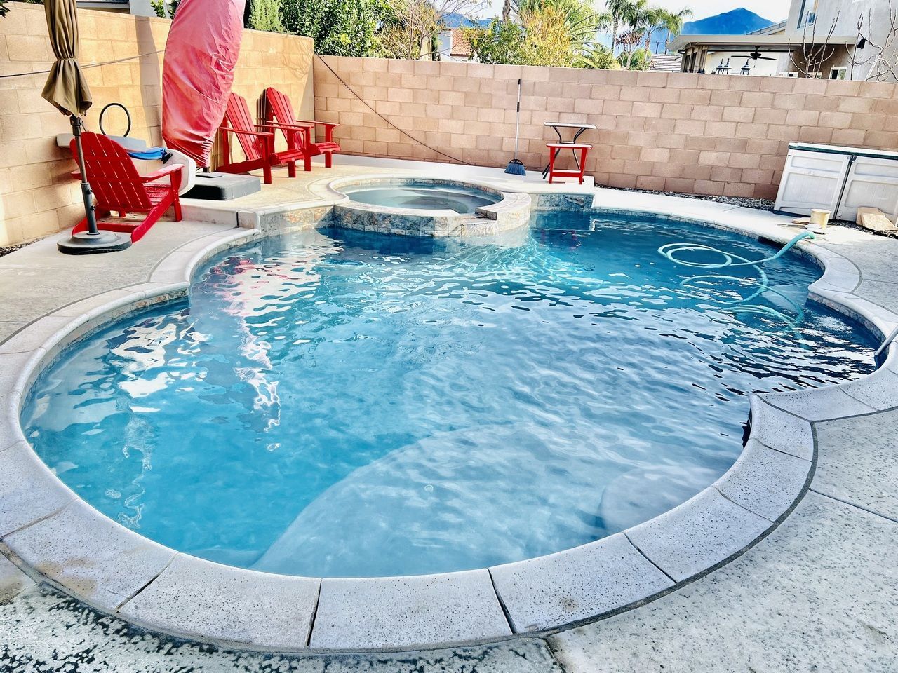 Pool with blue water and stone surround, red chairs, and a spa in a backyard setting.