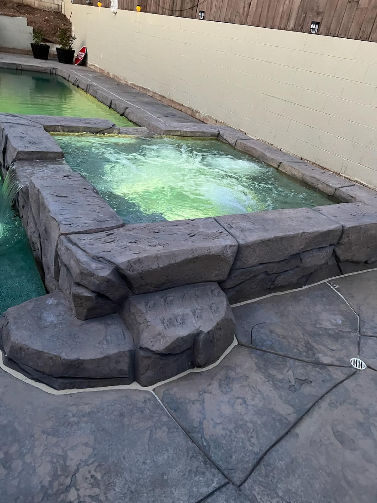 Stone-edged hot tub and pool. The hot tub is bubbling with illuminated green water. Concrete patio surrounding.