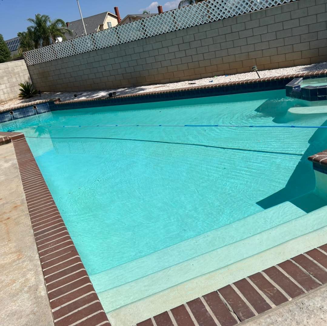 A rectangular swimming pool filled with turquoise water, with brick and concrete bordering, and a light-colored fence.