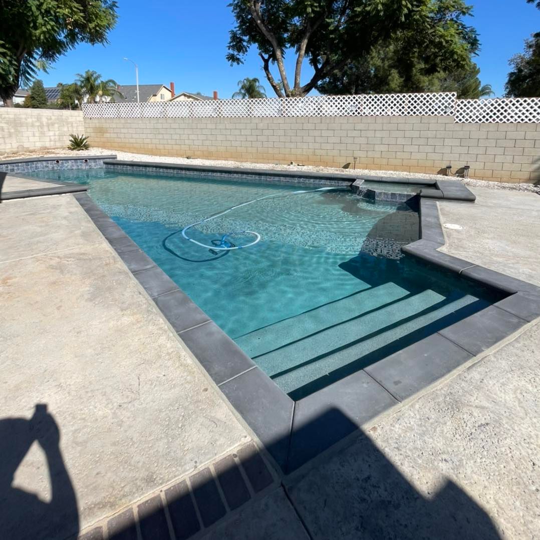 A rectangular swimming pool with steps, surrounded by concrete. Dark grey coping and a white wall in the background.