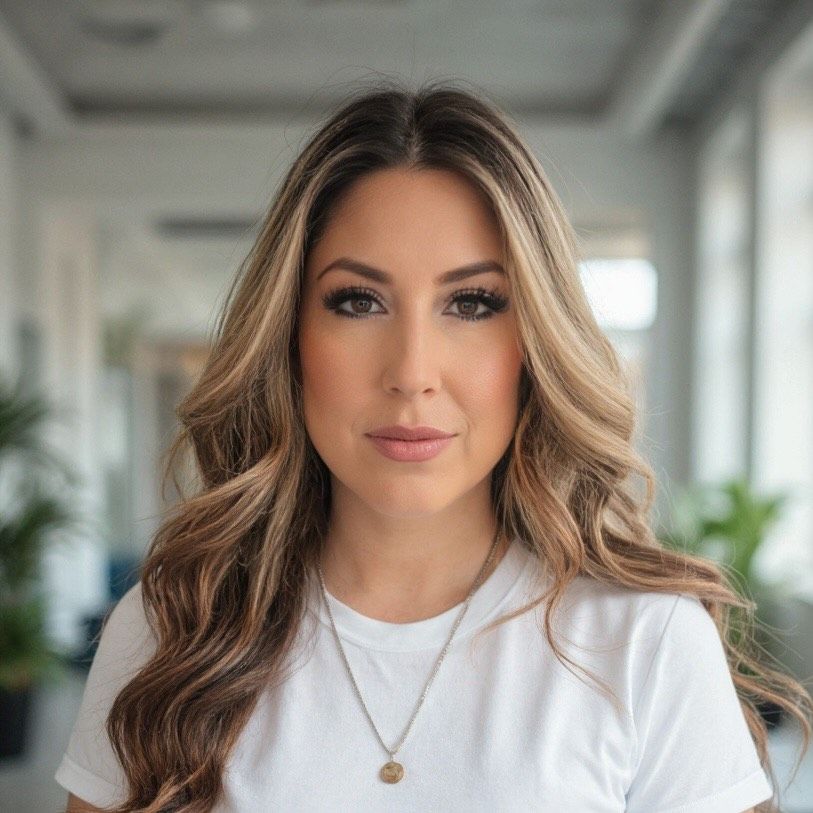 Woman with long wavy hair, wearing white shirt and necklace, in an office setting.