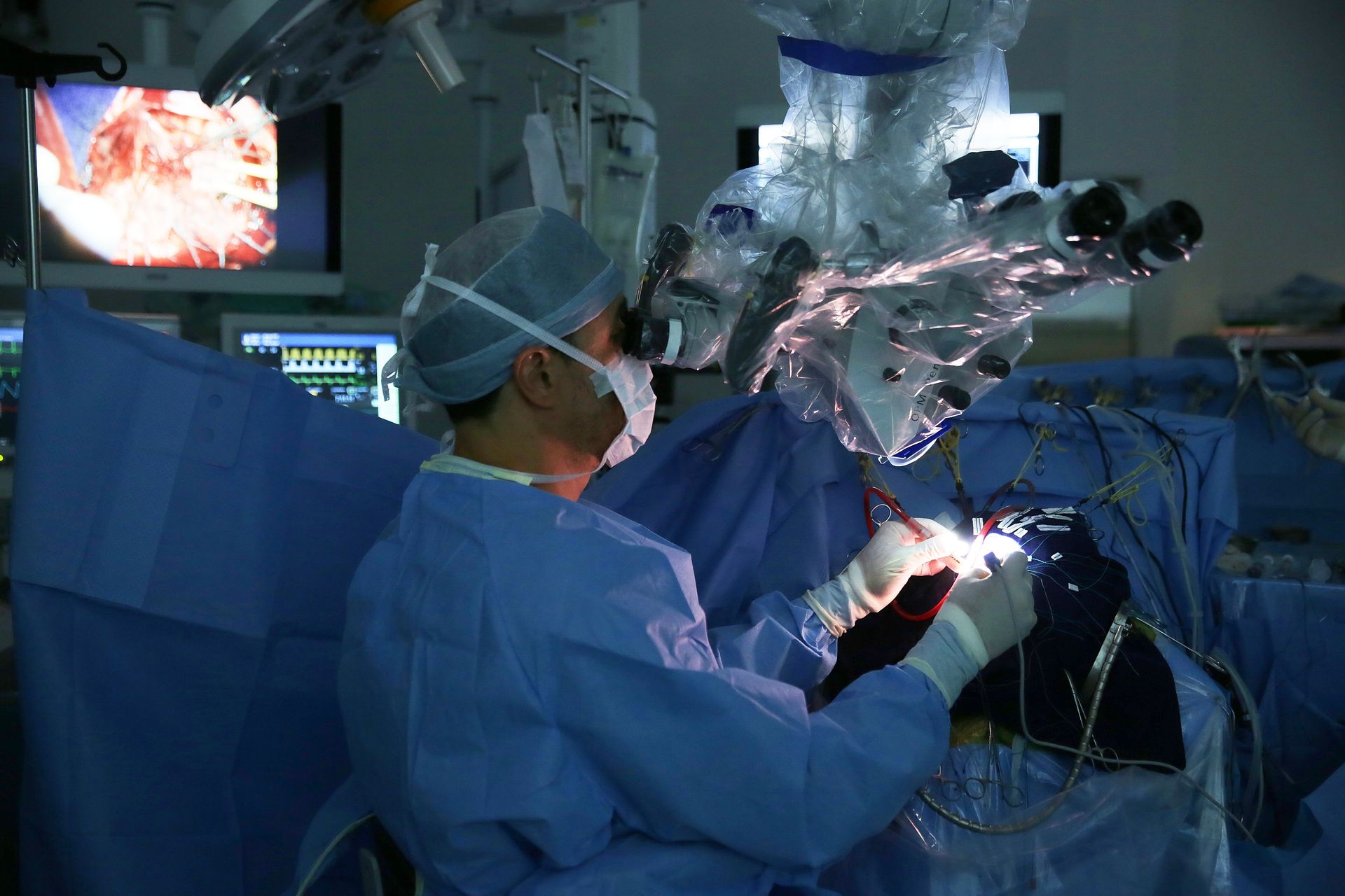 Cirurgião de uniforme azul e touca observando uma tomografia cerebral em um monitor em uma sala iluminada.