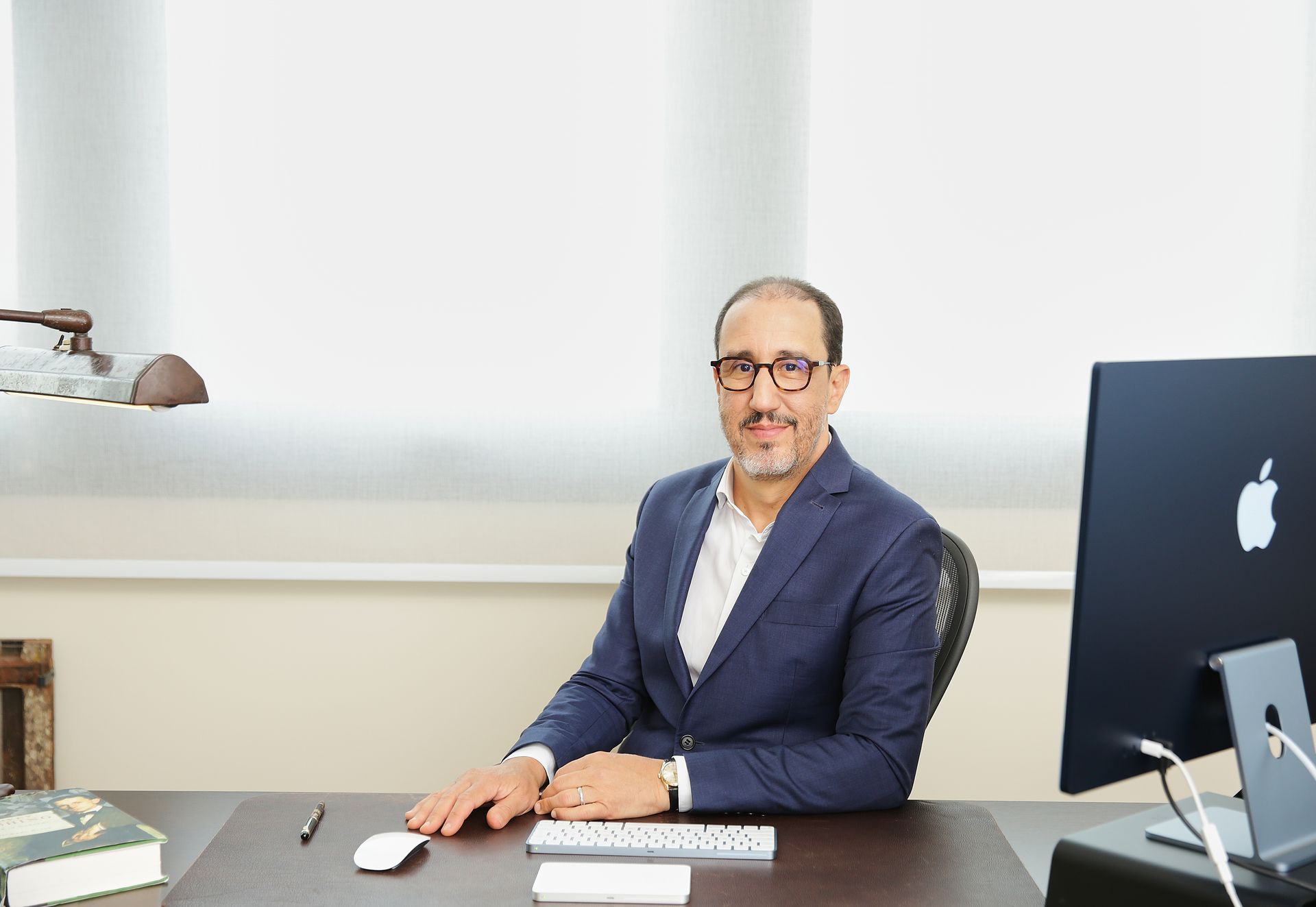 Homem de terno sentado à mesa, usando óculos, olhando para a câmera. Ambiente de escritório com computador e janela.
