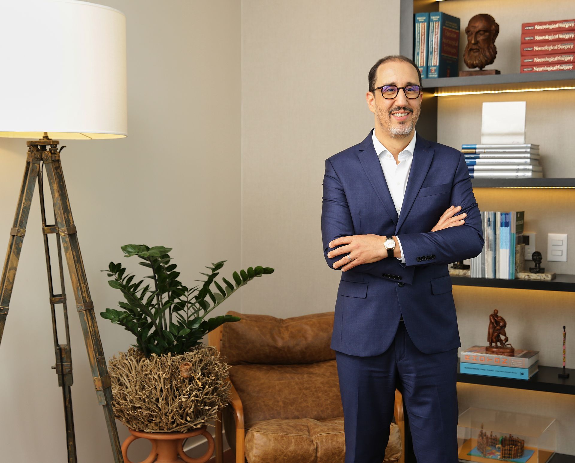 Homem de terno azul sorri, braços cruzados, em um escritório, ao lado de um abajur e uma estante.