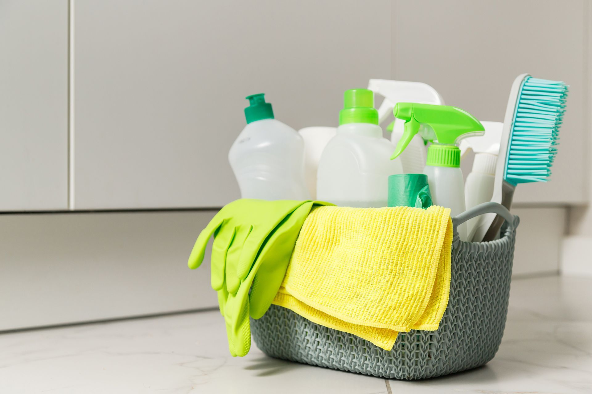 A gray basket filled with cleaning supplies: bottles of cleaner, gloves, a brush, and a yellow cloth, on a marble floor.