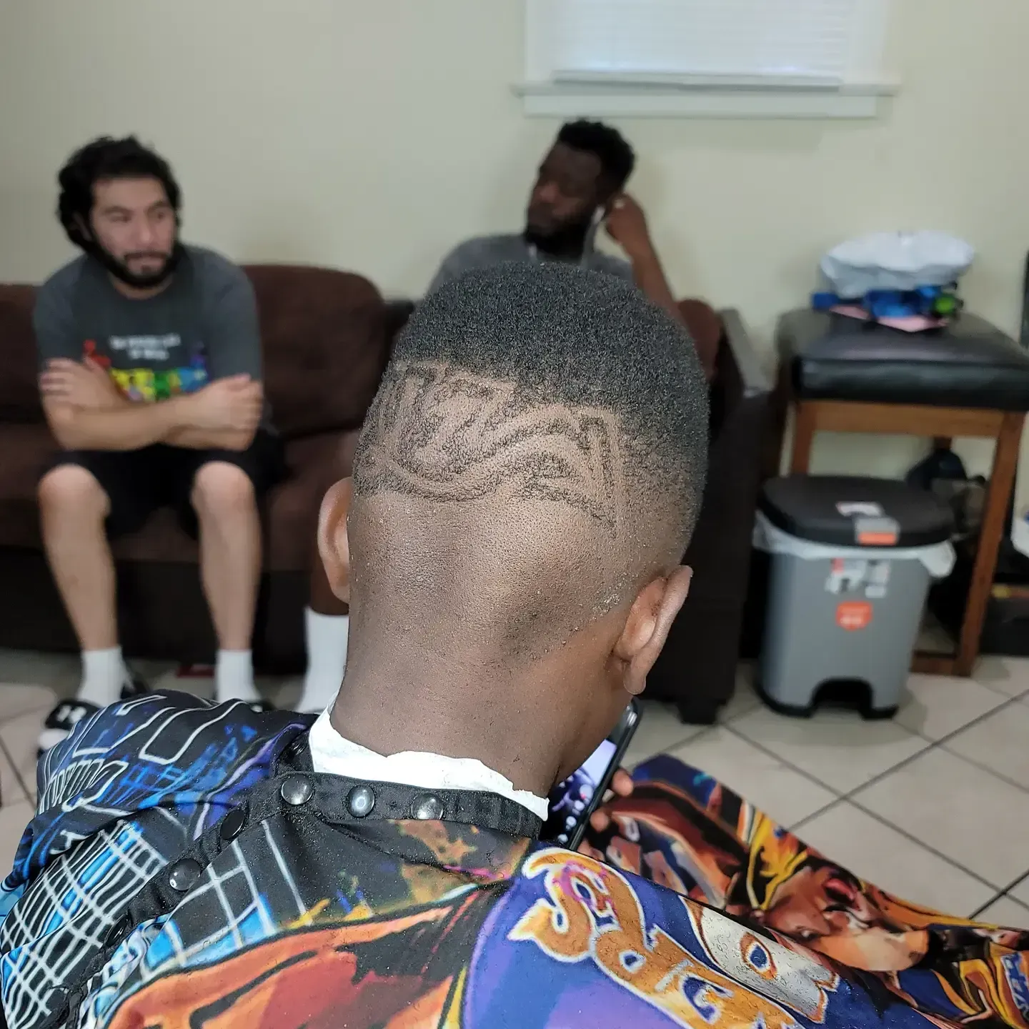 Two people sitting on a couch in a living room, viewed from behind someone wearing a colorful barber cape.