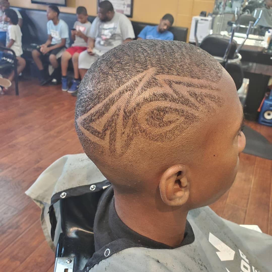 Boy with a geometric haircut in a barbershop, seated with people in the background.