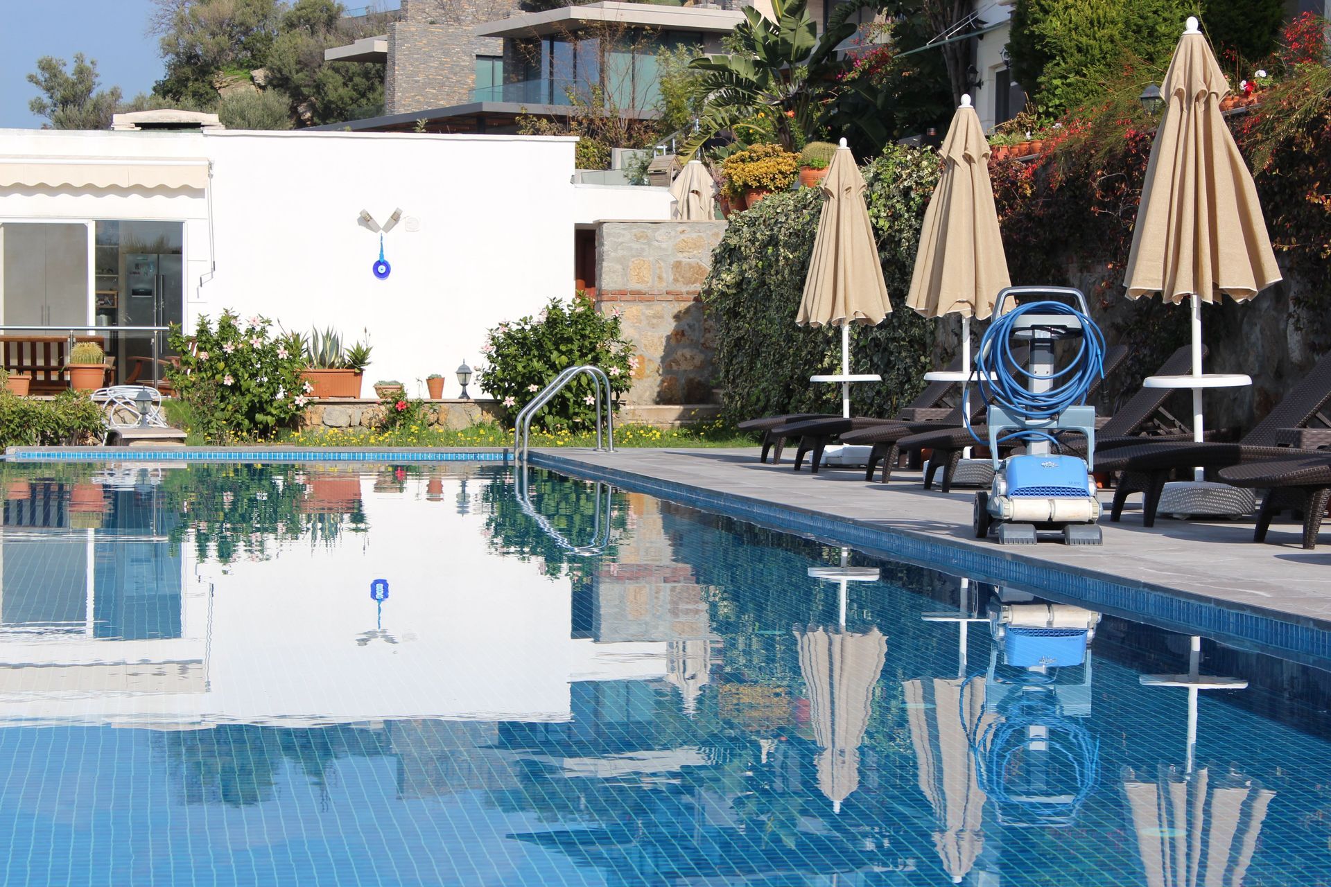 Outdoor pool with blue water, lounge chairs, closed umbrellas, and a pool-cleaning robot on the deck.