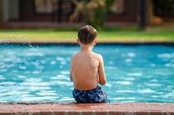 Child sitting by a blue swimming pool, facing the water, wearing blue swim trunks.