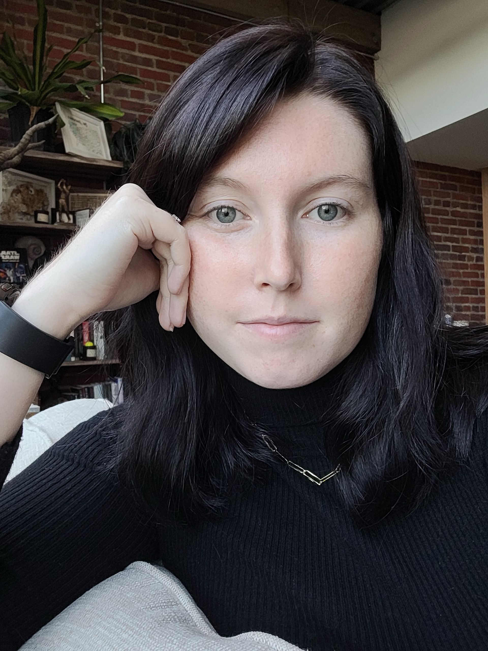 Woman with dark hair resting her head on her hand indoors, wearing a black sweater and wristband.