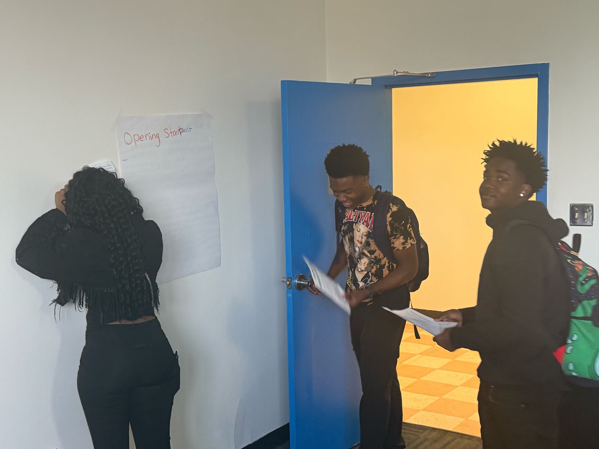Three people at a wall, one writing on paper, near an open blue door with bright light beyond.