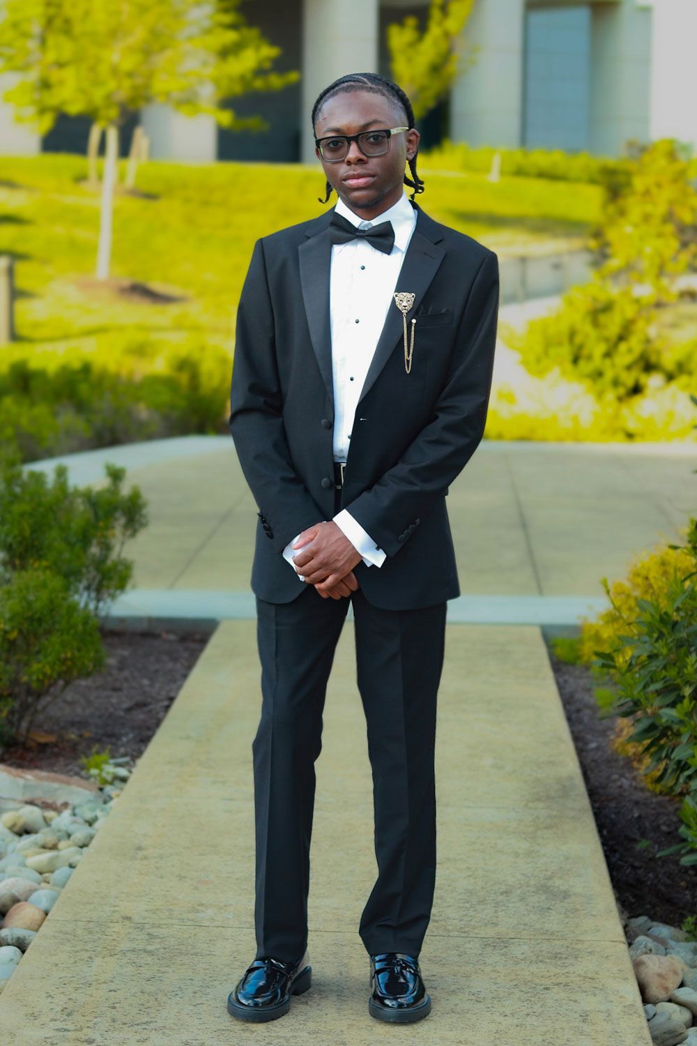 Person in a black tuxedo and glasses stands on a walkway in a landscaped garden.