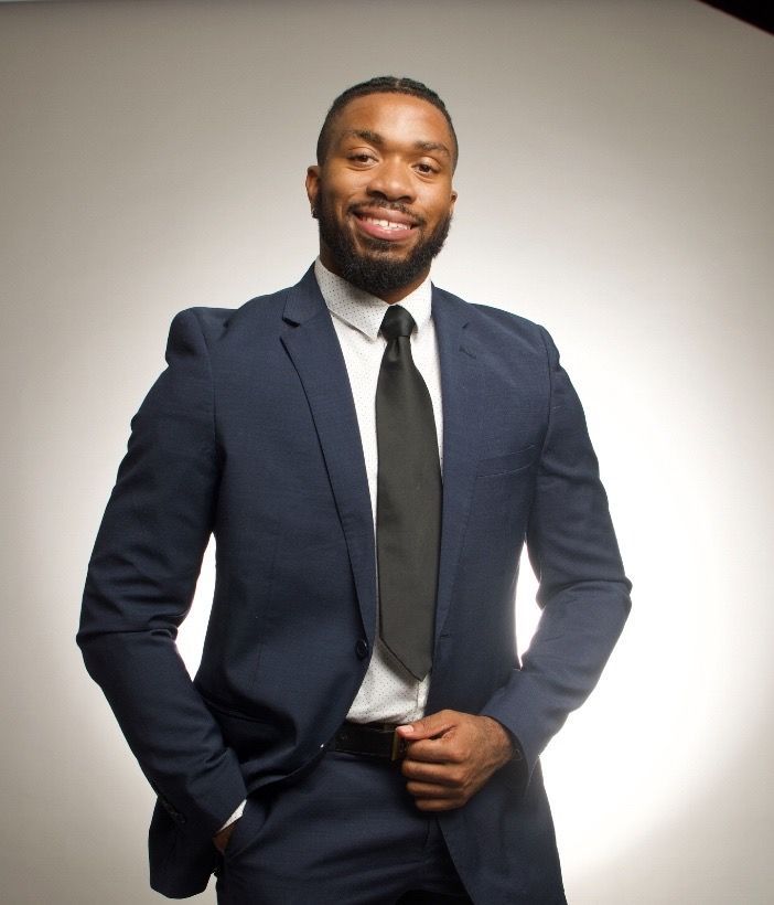 Man in navy suit, white shirt, and black tie, standing and smiling against a light gray background