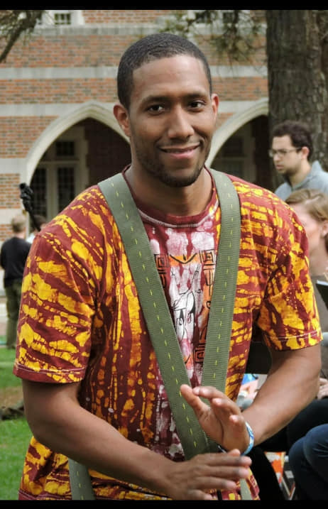 Person in a red-and-yellow patterned shirt standing outdoors, holding an instrument and smiling.