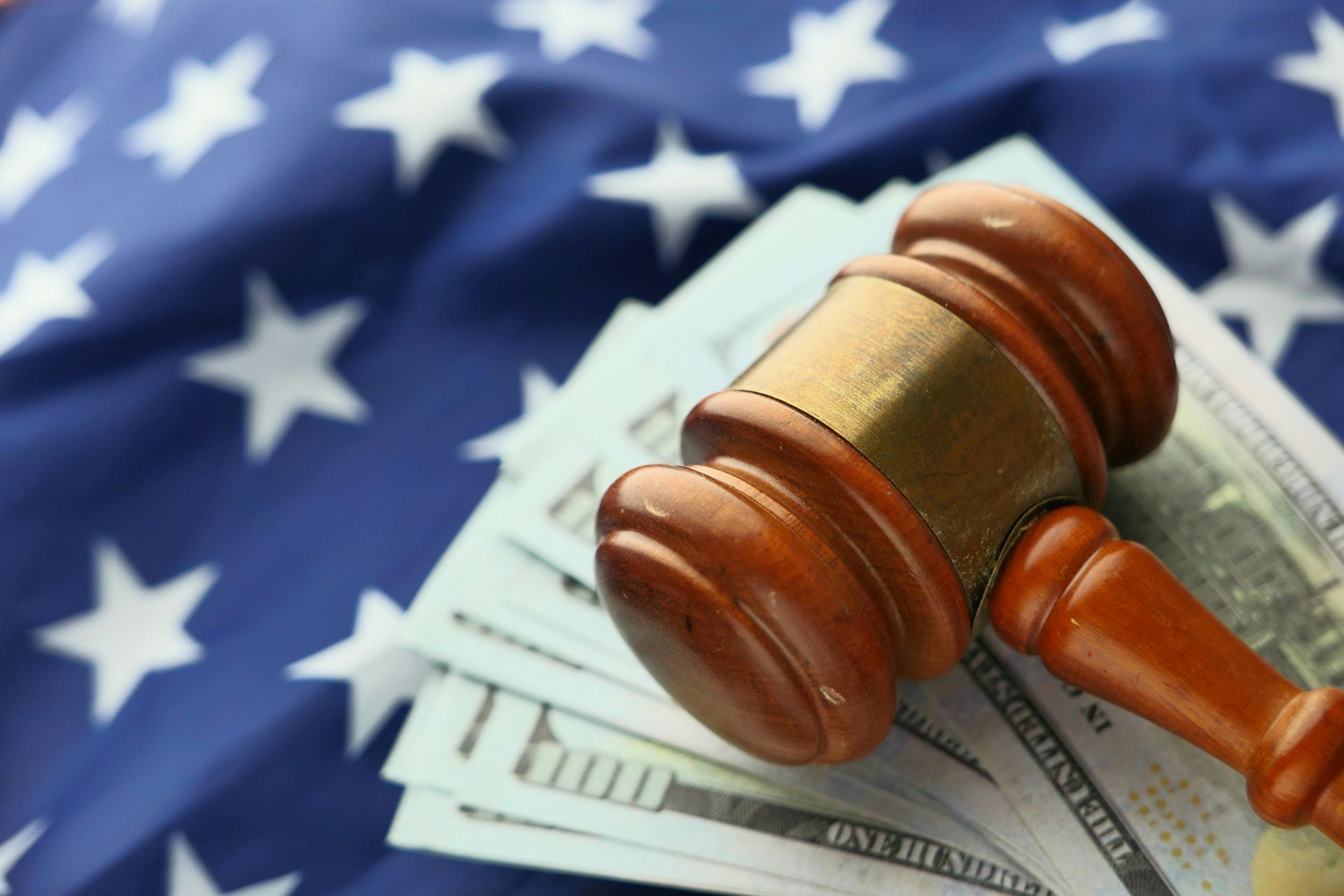 A wooden gavel resting on top of a stack of United States currency against a blurred American flag background.