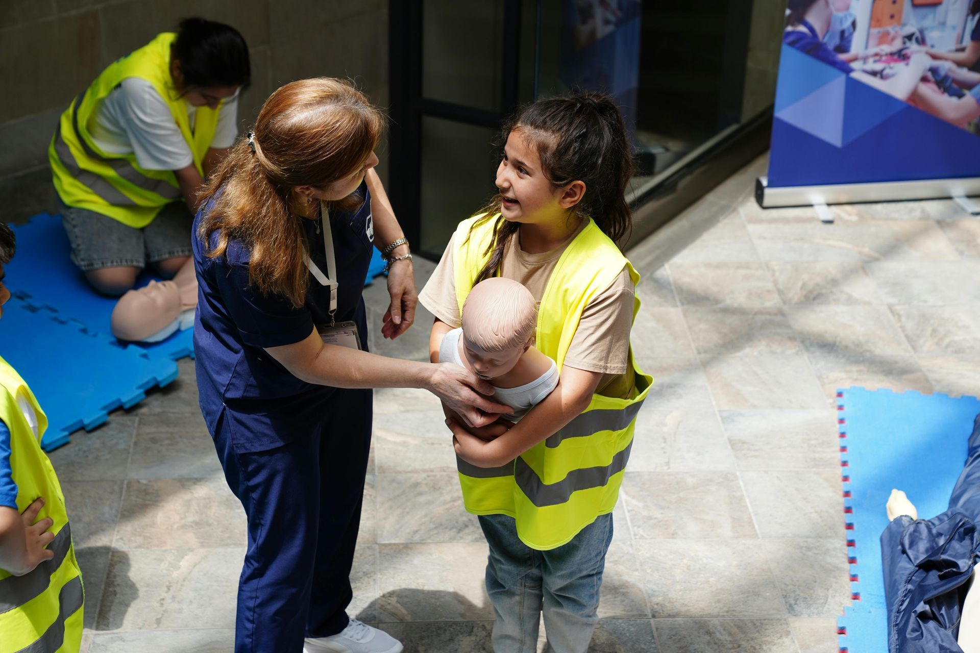 A healthcare professional guides a person wearing a yellow safety vest while they practice CPR techniques on a mannequin.