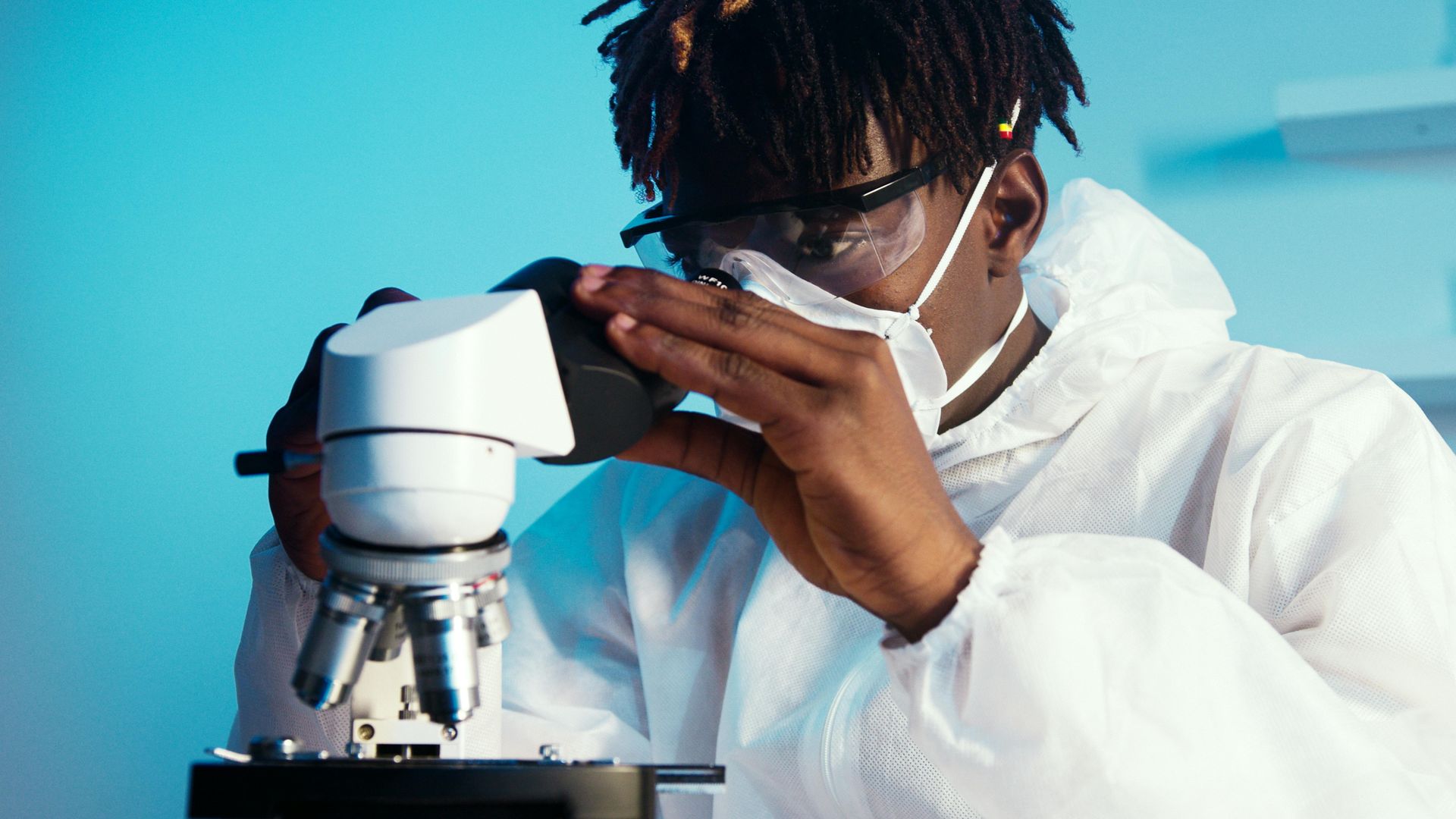 A scientist wearing a lab coat, mask, and safety glasses looks through the eyepiece of a microscope in a laboratory.