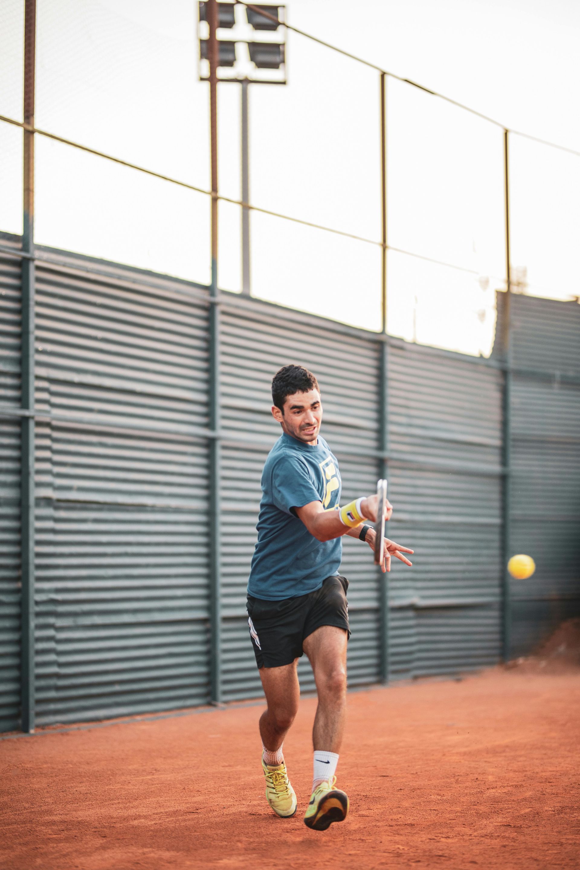 A tennis player in a blue t-shirt and dark shorts follows through on a forehand swing on a clay court.